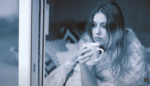 Woman looking out a window while holding a mug, shot through glass with cool blue color grading.