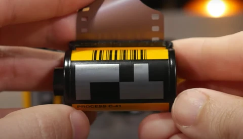 Close-up of a film canister being held in hands, showing the yellow barcode label and gray leader tape.