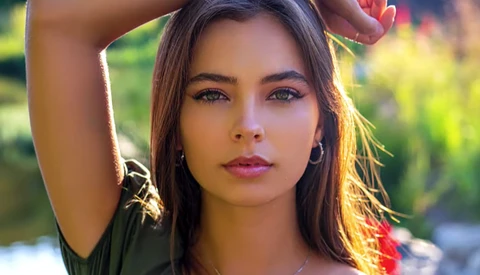 Portrait of a young woman with long dark hair in golden hour sunlight, hand raised to her head, looking directly at camera.