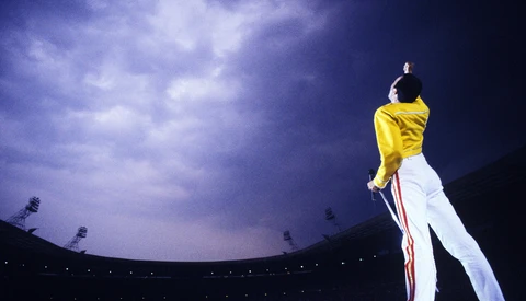 Performer on stage in yellow and white outfit against dramatic purple twilight sky and stadium lighting.