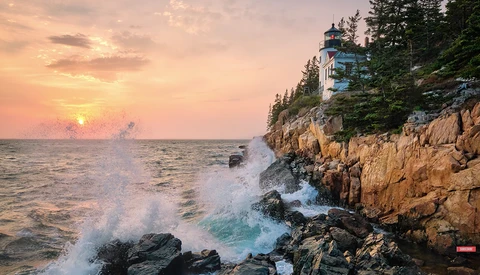 Crashing waves against rocky coastline at sunrise with lighthouse on forested cliff.