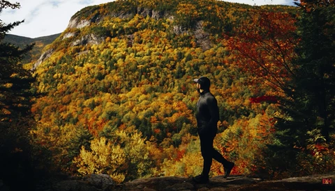 Person standing on a rocky outcrop facing a mountainside covered in vibrant autumn foliage.