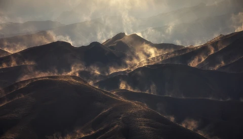 Wildfire smoke billows across mountainous terrain at golden hour, silhouetting ridgelines against dramatic clouds.