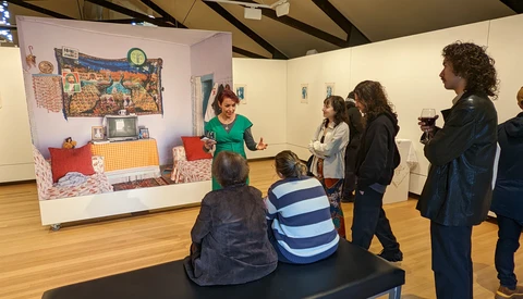 Gallery visitors observe an interactive bedroom display installation with a woman presenter in a green shirt explaining the exhibit.