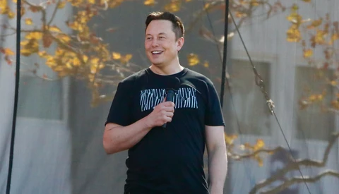Man in black t-shirt speaking with microphone against backdrop of golden autumn foliage.