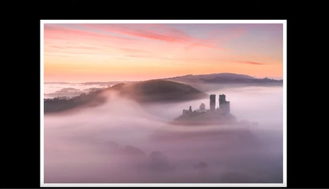 Medieval castle ruins emerging from thick fog at sunrise, with layered mountains visible in the distance.