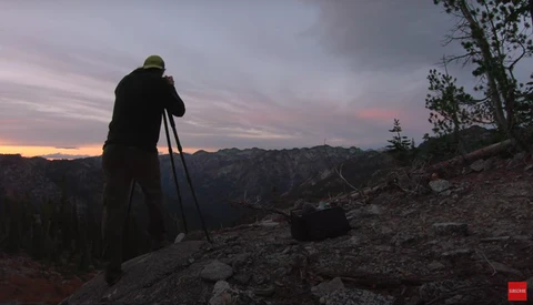 Photographer on a rocky hilltop aiming a telephoto lens toward a sunset landscape.