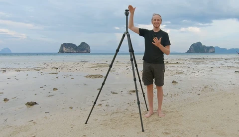 Photographer with tripod on beach gestures toward camera during golden hour shoot.