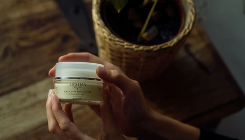 Hand holding a cosmetic jar labeled Lesina Black Star Moisturizer against a wooden surface with woven basket backdrop.
