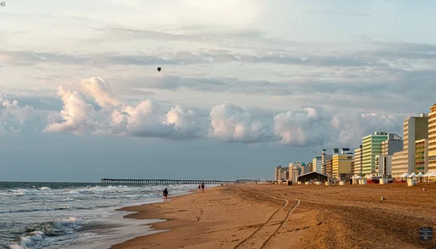 Beach scene at sunset with high-rise hotels lining the shore, a wooden pier extending into calm waters, and dramatic cloud formations on the horizon.