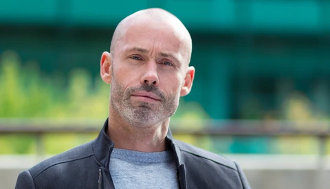 Bald man with facial hair wearing a dark jacket over a gray shirt, photographed outdoors with blurred green background.
