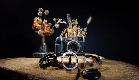Rangefinder camera with brass lens rings and dried flowers arranged on weathered wood against black background.