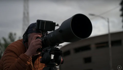 Photographer holding a telephoto lens and camera mounted on a tripod, composing a shot outdoors under overcast skies.