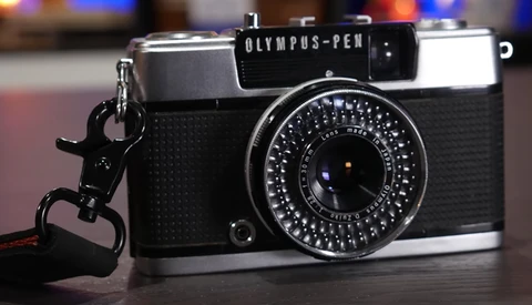 Olympus PEN half-frame camera with rangefinder lens and wrist strap, displayed on dark surface with blurred background lights.