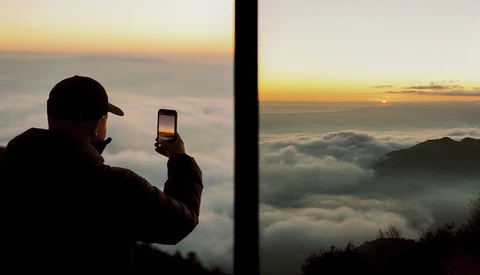 Silhouetted figure photographing a sunrise above clouds with a smartphone.