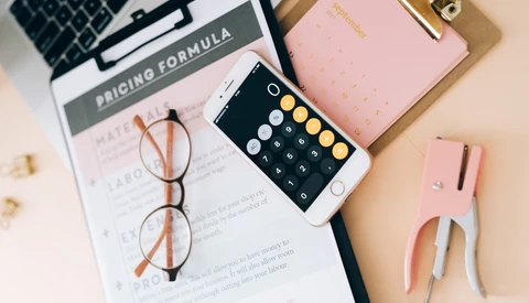 Flat lay workspace with laptop, smartphone showing calculator app, eyeglasses, pink notebook, and printed documents on beige surface.