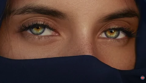 Close-up portrait of a person's eyes with golden-amber irises and dark brows, lower face obscured by dark fabric.