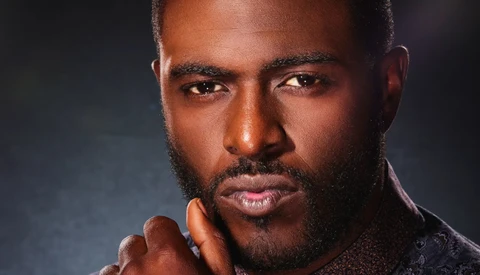 Man with beard gazing directly at camera, hand raised to face in thoughtful pose.