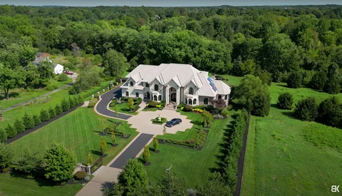 Aerial view of a large two-story stone and brick residence with curved driveway on manicured grounds surrounded by mature trees.