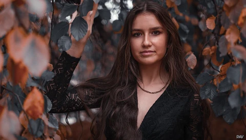 Portrait of a woman with long dark hair framed by autumn foliage in warm orange and brown tones.