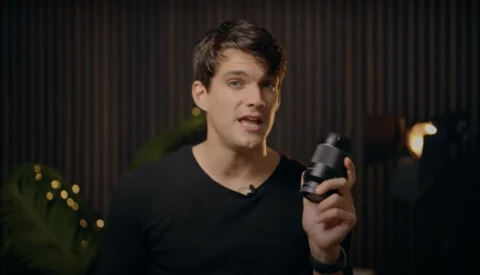 Man in black v-neck shirt holding a microphone while speaking, shot against a dark corrugated backdrop.