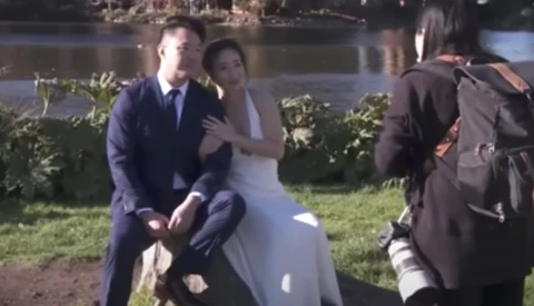 Wedding photographer shooting bride and groom posing by waterside during outdoor ceremony.