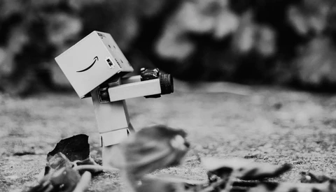 White box character with Amazon smile logo tilting head while holding camera on rocky ground.