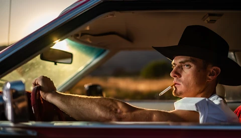 Man in cowboy hat smoking cigarette while driving classic car at sunset.