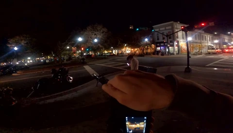 Nighttime street scene shot from a bicycle handlebar perspective, showing an urban downtown area with illuminated storefronts and street lighting.
