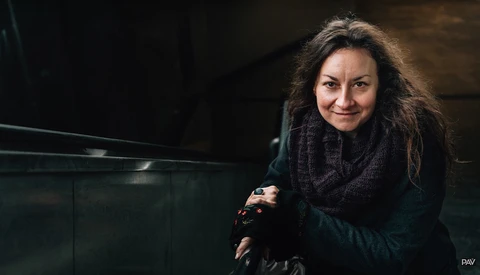 Woman with long dark hair wearing a black scarf and jacket, smiling at the camera in an indoor setting.