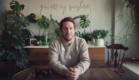 Man seated at wooden table with camera, surrounded by potted plants and natural window light.