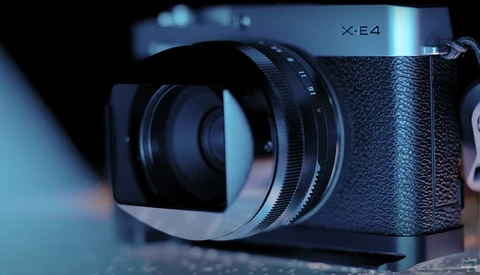Fujifilm X-E4 mirrorless camera with attached lens hood, shown in three-quarter view against a soft-focused blue background.