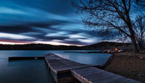 Wooden dock extending into a still lake at dusk, with bare trees and distant shoreline beneath a dramatic twilight sky.