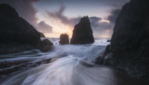 Sea stacks silhouetted against a twilight sky with waves washing across the foreground beach.