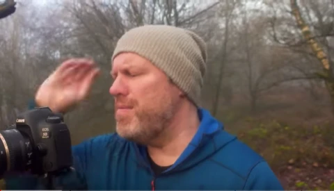 Photographer holding a camera with lens shade, wearing beanie and blue jacket in bare winter woods.
