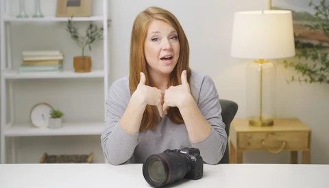 Woman with reddish-brown hair gesturing enthusiastically toward a professional DSLR camera on a white desk.