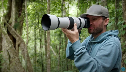 Photographer with telephoto lens in forest setting, wearing gray cap and blue jacket.