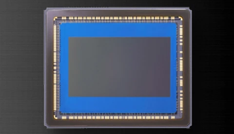 Close-up photograph of a camera image sensor die mounted in a clear plastic package with gold contact pins around the perimeter.