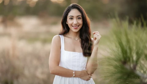 Woman in white dress smiling at camera in outdoor setting with blurred natural background.