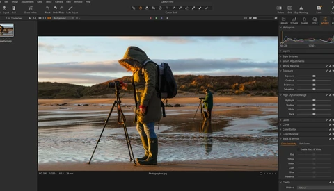 Photographer in yellow jacket operating tripod-mounted camera on beach at golden hour with two figures in distance.