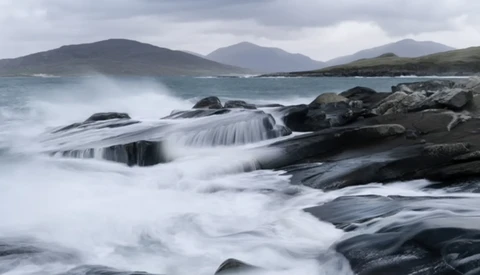 Waves crash over dark rocks along a rocky coastline with mountains in the distant background.