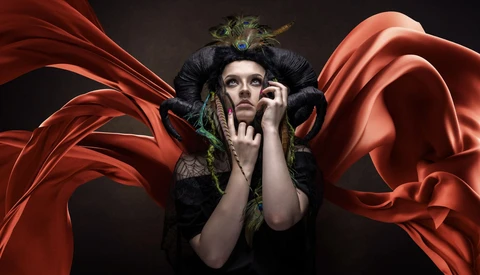 Person in dark costume with feathered headdress and dramatic red fabric wings against black background.