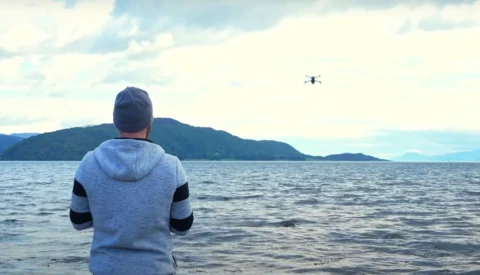 Person in blue and black hoodie standing in shallow water, operating a drone hovering over the ocean.