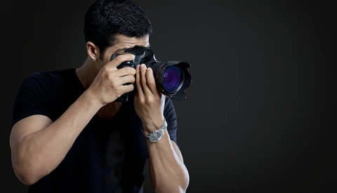 Male photographer holding a DSLR camera with telephoto lens, positioned against dark background.