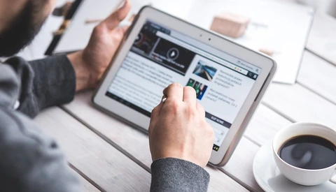 Person using tablet to view a website with video content and articles at a desk workspace.