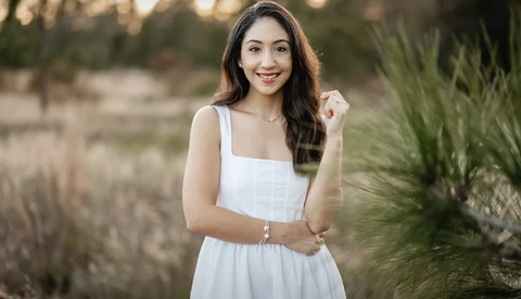 Woman in white sleeveless dress posing outdoors during golden hour with natural vegetation in background.