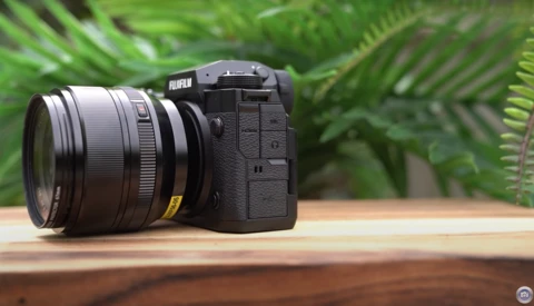 Fujifilm mirrorless camera with attached lens on wooden surface, green foliage in background.