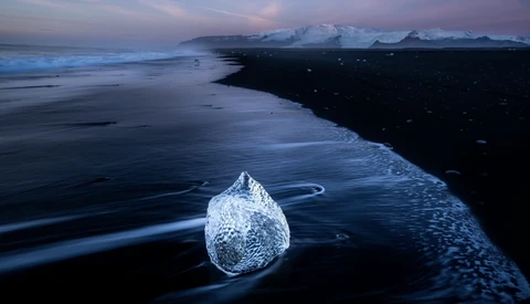 Translucent ice chunk on dark volcanic sand beach during twilight hour.