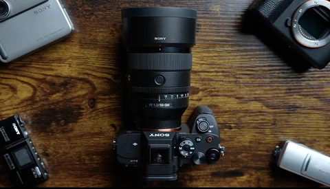 Sony camera body with attached telephoto lens surrounded by additional lenses and camera equipment on wooden surface.