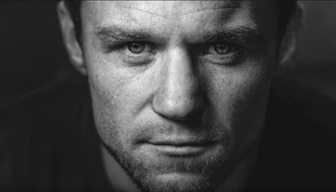 Close-up black and white portrait of a man with intense gaze and stubbled jaw against dark background.